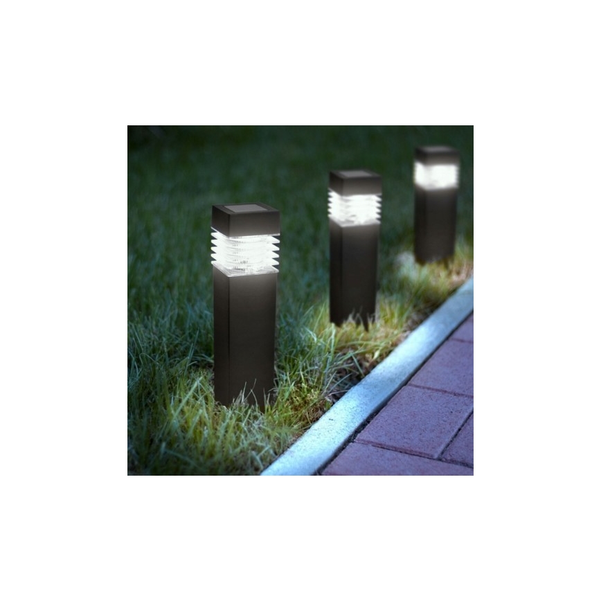 Tri záhradné solárne lampy svietia v tráve popri chodníku. Lampa je vysoká, štvorcová, tmavej farby a svieti teplým svetlom.