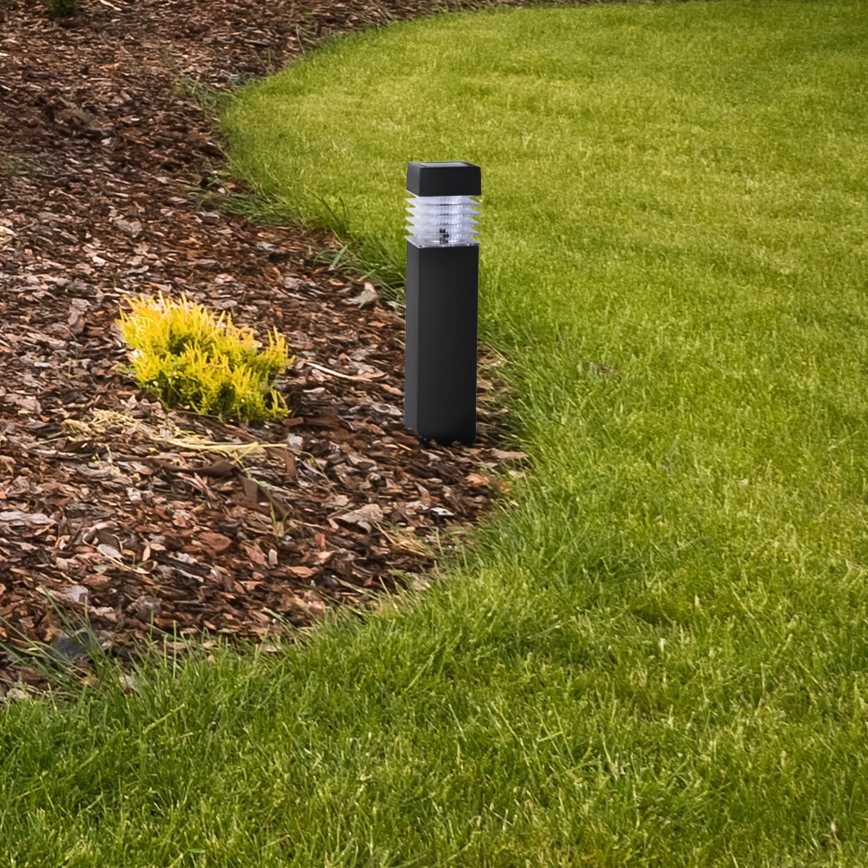 Záhradné svietidlo stojí v trávniku. Svietidlo je čierne, štvorcového tvaru, zapustené v mulči. Okolo je krátko strihaný trávnik a hnedý drevený mulč.