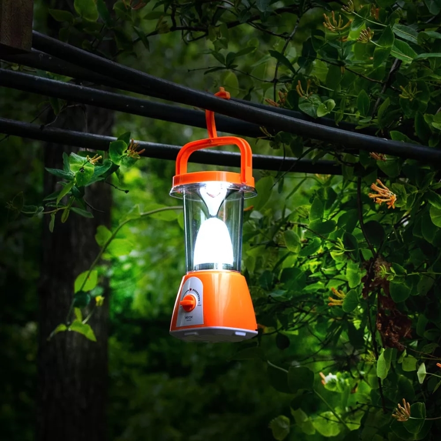 Oranžová prenosná lampa svieti. Je zavesená na elektrických kábloch v lesnom prostredí medzi zelenými listami.