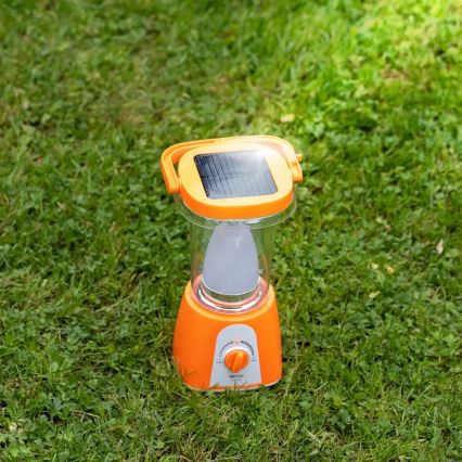 Solárna lampa stojí na tráve. Je oranžovej farby, má integrovaný solárny panel a LED svetlo. Na spodku je otočný prepínač s nápisom Veron.