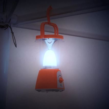 Oranžová prenosná lampa svieti. Je zavesená na háku pod stropom. Lampa má otočný ovládač s nápisom „VOL“.