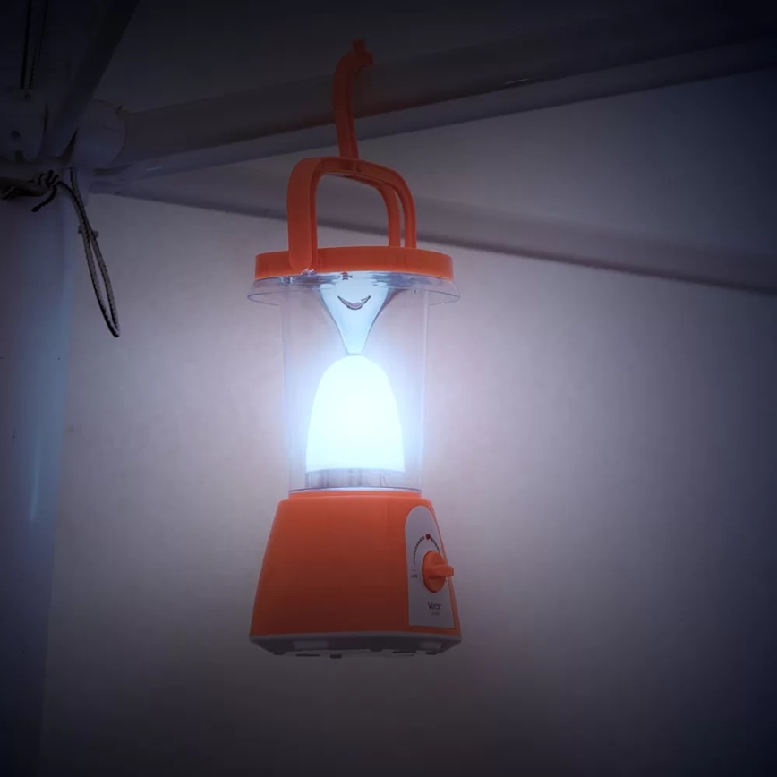 Oranžová prenosná lampa svieti. Je zavesená na háku pod stropom. Lampa má otočný ovládač s nápisom „VOL“.
