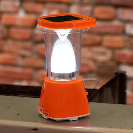 Solárna lampa svieti. Je oranžovej farby, s priehľadnou nádobkou a LED svetlom. Stojí na kovovej ploche pred tehlovou stenou.