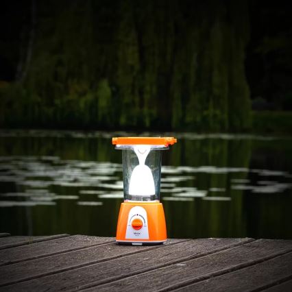Prenositelná oranžová lampa svieti na drevenom móle pri jazere. Svetlo pochádza z vnútornej žiarovky. Na spodku lampy je viditeľný nápis „Verox“.