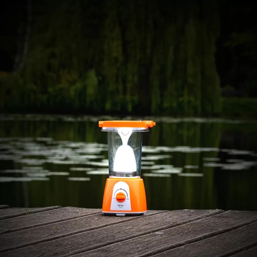 Prenositelná oranžová lampa svieti na drevenom móle pri jazere. Svetlo pochádza z vnútornej žiarovky. Na spodku lampy je viditeľný nápis „Verox“.