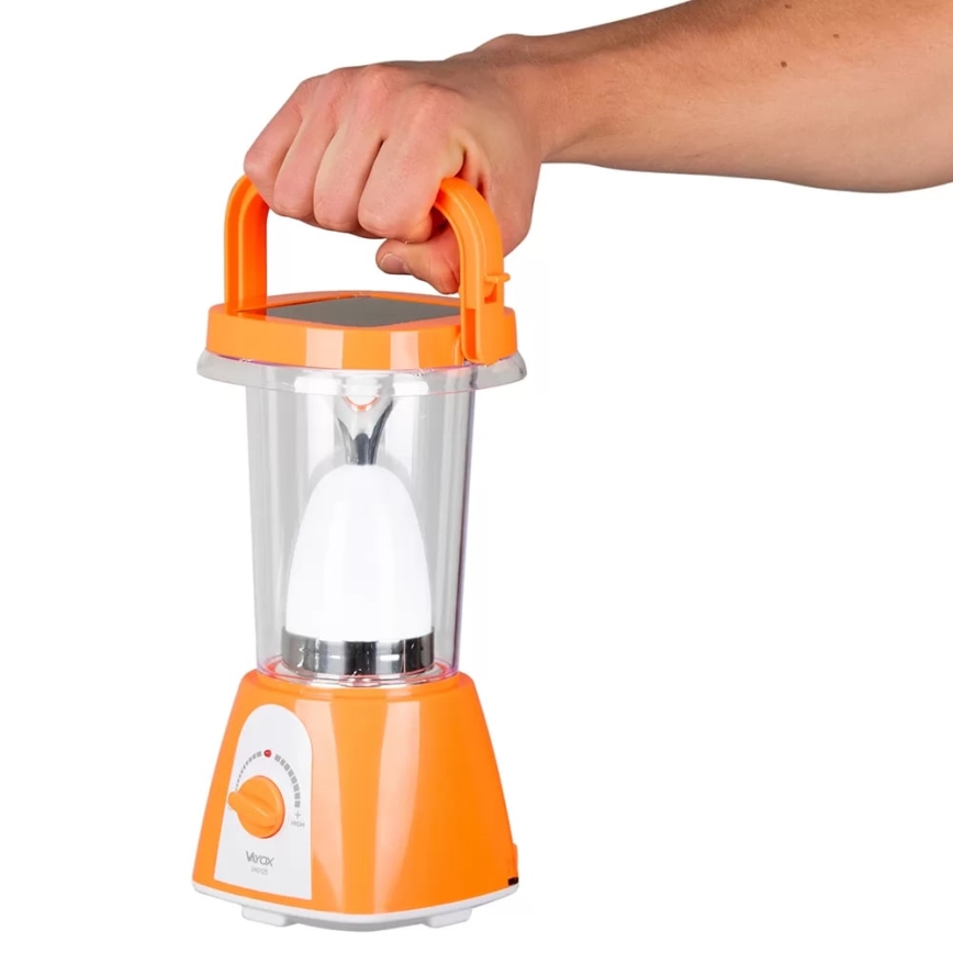 Ruka drží oranžovú solárnu lampu s priehľadným telom a bielym svetelným zdrojom. Lampa má horné držadlo a otočný spínač s nápisom Virax. Biela plocha na vrchu lampy naznačuje solárny panel.