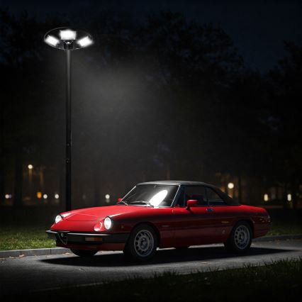 Červené Alfa Romeo Spider stojí zaparkované v noci pri pouličnom osvetlení. Auto je osvetlené lampou. V pozadí sú stromy a slabo viditeľné ďalšie svetlá.