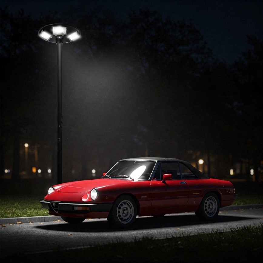 Červené Alfa Romeo Spider stojí zaparkované v noci pri pouličnom osvetlení. Auto je osvetlené lampou. V pozadí sú stromy a slabo viditeľné ďalšie svetlá.