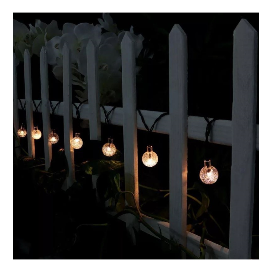 Svietiace guľôčky svietia v noci. Sú pripevnené na šnúre, ktorá je prevesená cez biely plot. Rastlinstvo je viditeľné pod plotom.