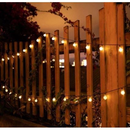 Guľôčkové svetielka s teplým svetlom sú omotané okolo dreveného plota vonku v noci. Svietiace guľôčky sú rozmiestnené pozdĺž tenkého kábla.