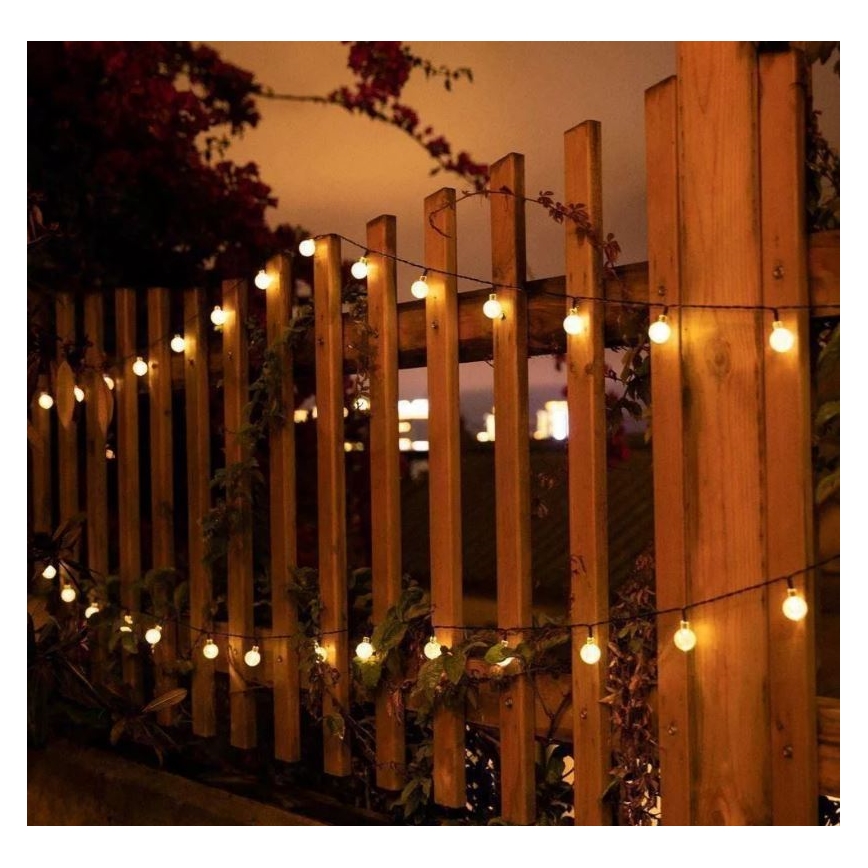 Guľôčkové svetielka s teplým svetlom sú omotané okolo dreveného plota vonku v noci. Svietiace guľôčky sú rozmiestnené pozdĺž tenkého kábla.