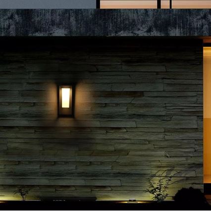 Vonkajšie nástenné svietidlo svieti na kamennej stene. Svietidlo je obdĺžnikového tvaru, zapustené do steny. V okolí je tma a vidieť nízku zeleň.