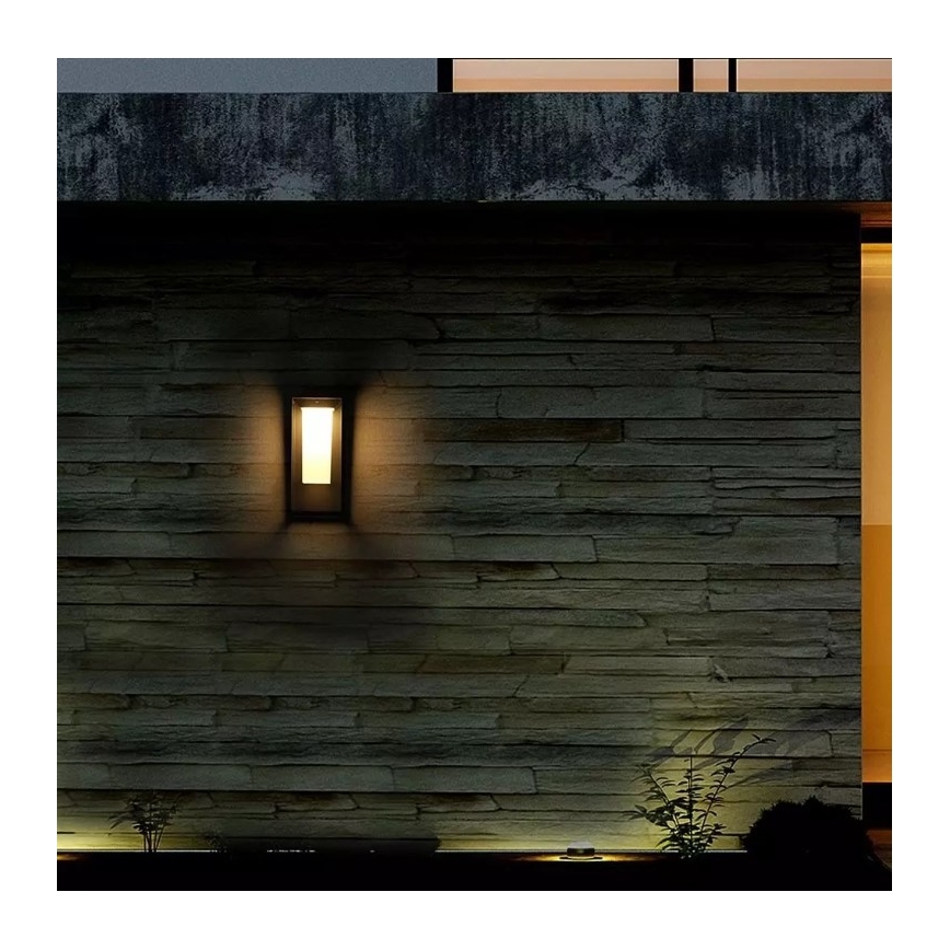 Vonkajšie nástenné svietidlo svieti na kamennej stene. Svietidlo je obdĺžnikového tvaru, zapustené do steny. V okolí je tma a vidieť nízku zeleň.