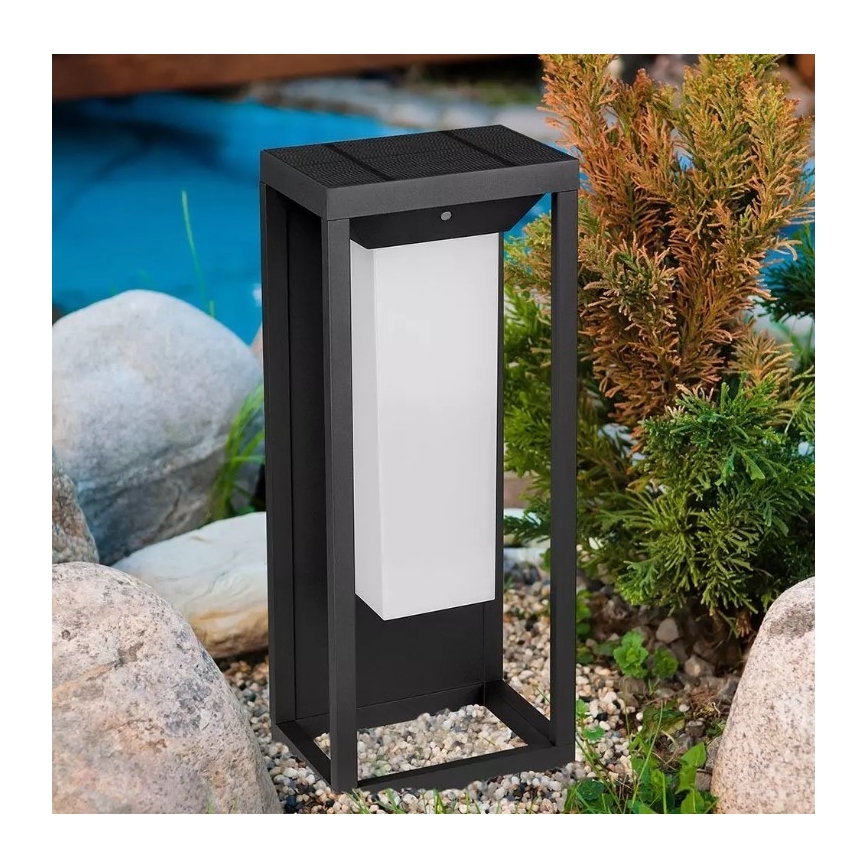 Solárna záhradná lampa stojí na kamenistej pôde medzi skalami a kríkmi. Lampa svieti bielym svetlom. Je obdĺžniková, tmavosivá s integrovaným solárnym panelom.