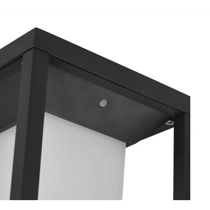 Čierny kovový stojací svietič s bielym difúzorom. Svietič je umiestnený vo vonkajšom priestore. Na vrchu je viditeľné malé tlačidlo.