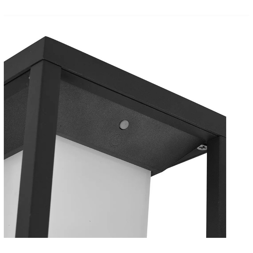 Čierny kovový stojací svietič s bielym difúzorom. Svietič je umiestnený vo vonkajšom priestore. Na vrchu je viditeľné malé tlačidlo.