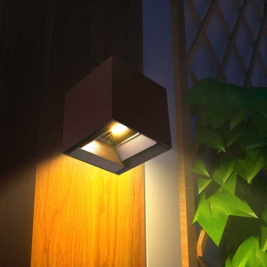 Kubická nástenná lampa svieti. Je umiestnená na drevenom povrchu vedľa zelenej rastliny a drevenej mriežky.