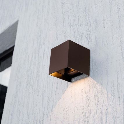 Kubická vonkajšia nástenná lampa svieti na svetlohnedú omietkovú stenu. Lampa je tmavohnedá, pripevnená k stene vedľa okna.