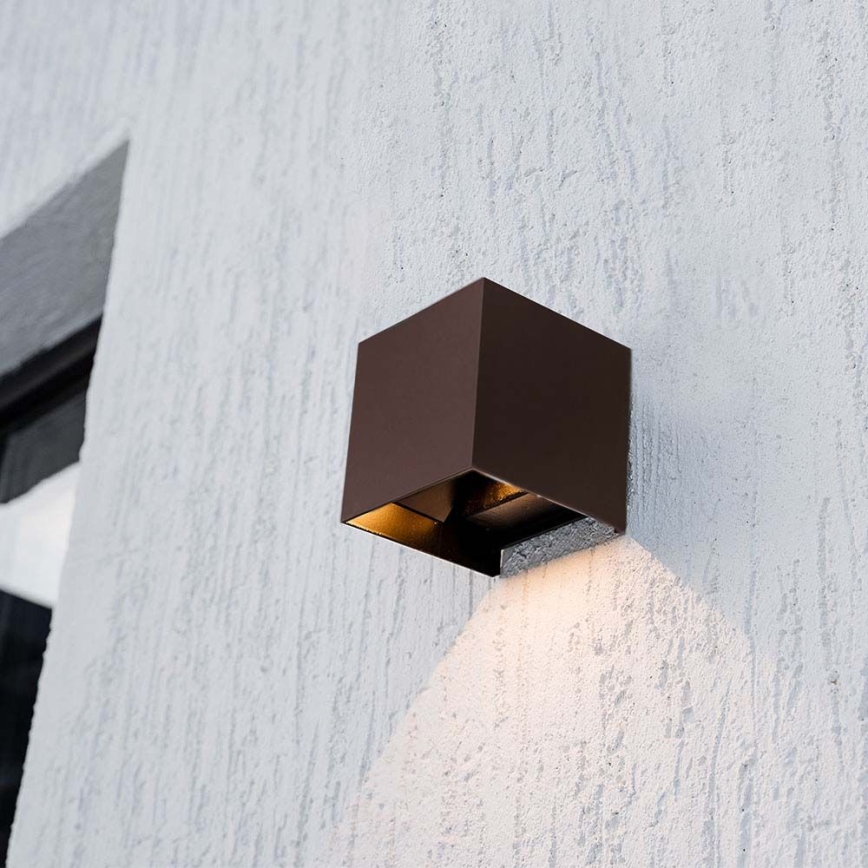 Kubická vonkajšia nástenná lampa svieti na svetlohnedú omietkovú stenu. Lampa je tmavohnedá, pripevnená k stene vedľa okna.