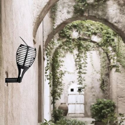 Solárna lampa sa montuje na stenu starobylého, zalesneného nádvoria. Lampa má tvar fakle s čiernym kovovým krytom. Na pozadí sú kamenné oblúky a dvere.