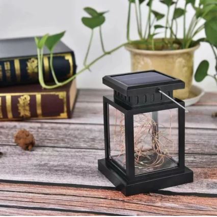 Solárna lampa s LED osvetlením stojí na drevenom povrchu. Je obdĺžnikového tvaru s priehľadnými stranami, v ktorých sú viditeľné svetelné reťazce. Na vrchu je solárny panel. V pozadí sú knihy a rastlina v kvetináči.