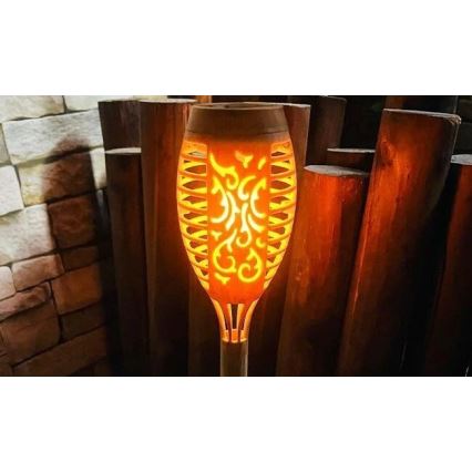 LED lampa tvaru fakle svieti teplým oranžovým svetlom. Lampa je umiestnená pred dreveným plotom. Svetlo simuluje plameň s vyrezávaným vzorom.