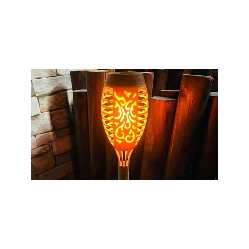 LED lampa tvaru fakle svieti teplým oranžovým svetlom. Lampa je umiestnená pred dreveným plotom. Svetlo simuluje plameň s vyrezávaným vzorom.