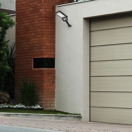 Vonkajšie nástenné LED svetlo je pripevnené k stene domu, vedľa garážových dverí. Svetlo je orientované smerom nadol. Dom má červenohnedú tehlovú stenu a svetlohnedé garážové dvere.