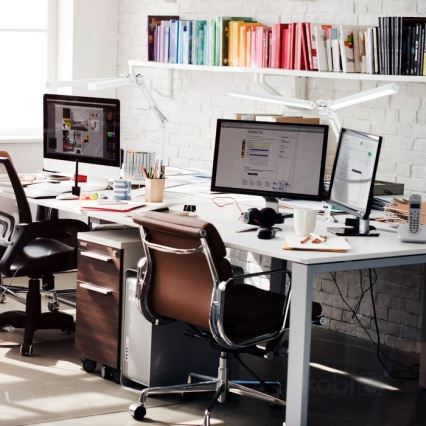 Dva počítače stoja na veľkom bielom stole v kancelárii. Stolička z hnedého koženého materiálu sa nachádza oproti monitorom. Stenu zdobia police s knihami.