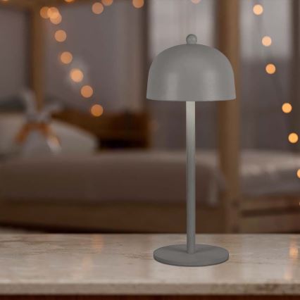 Stolová lampa s kupolou stojí na svetlom povrchu. Je sivá a má jednoduchý dizajn. Pozadie je rozmazané a zobrazuje spálňu so svetelnými girlandami.