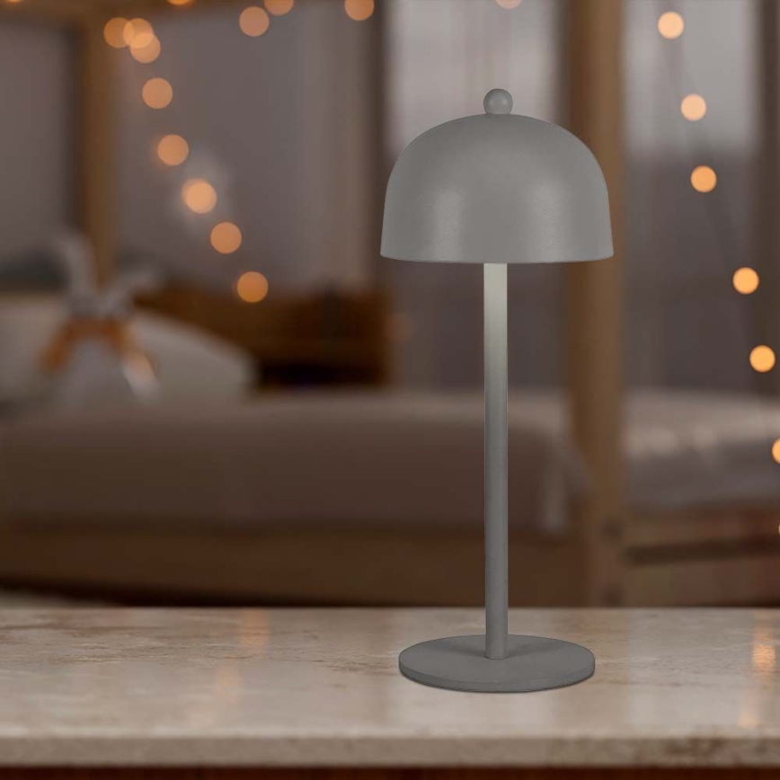 Stolová lampa s kupolou stojí na svetlom povrchu. Je sivá a má jednoduchý dizajn. Pozadie je rozmazané a zobrazuje spálňu so svetelnými girlandami.