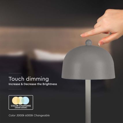 Šedá stolová lampa s kupolovitým tienidlom. Prst sa dotýka vrcholu tienidla, čím sa reguluje jas. Text uvádza: „Touch dimming. Increase & Decrease the Brightness. COLOR CHANGING (3000K-6000K). Color 3000k-6000k Changeable.“