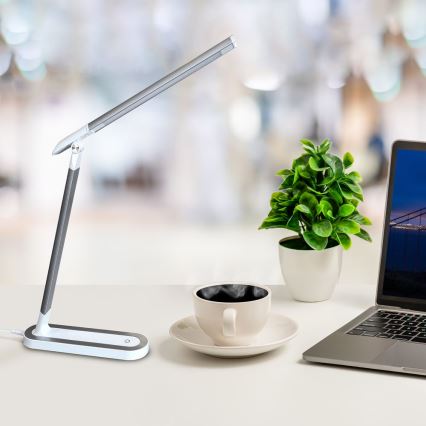 Stolná lampa s LED osvetlením je umiestnená na bielom stole vedľa šálky kávy, kvetináča a notebooku. Lampa je šedo-biela a má dlhý, tenký ramenný výložník. Pozadie je rozostrené a znázorňuje kancelársky priestor.