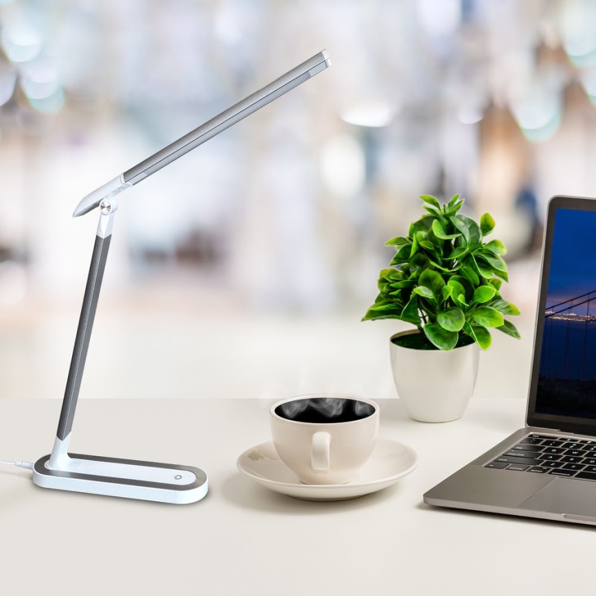 Stolná lampa s LED osvetlením je umiestnená na bielom stole vedľa šálky kávy, kvetináča a notebooku. Lampa je šedo-biela a má dlhý, tenký ramenný výložník. Pozadie je rozostrené a znázorňuje kancelársky priestor.