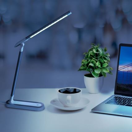 Stolová lampa s LED svetlom svieti na bielom stole. Na stole je tiež šálka kávy, črepníková rastlinka a notebook s obrázkom mosta na obrazovke.