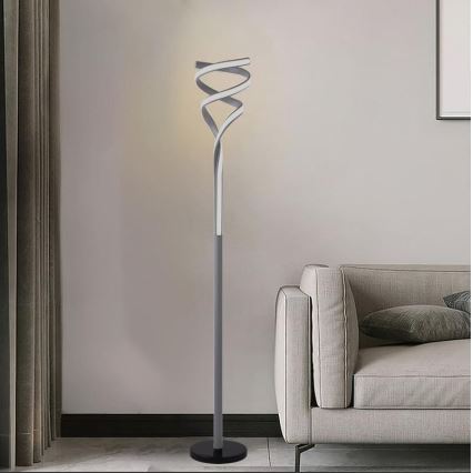 Stojacia lampa svieti v obývacej izbe. Jej tienidlo je špirálovito tvarované, sivé, na tmavošedom stojane. Nachádza sa blízko svetlej pohovky.