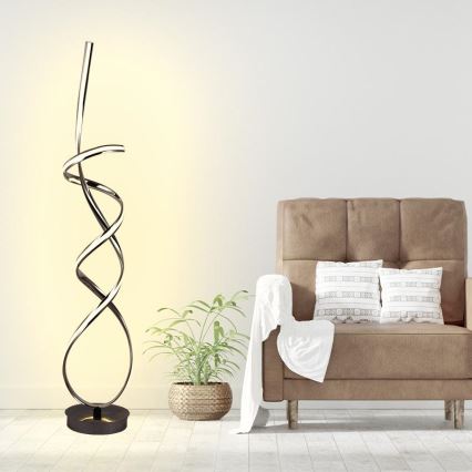 Stojacia lampa s LED osvetlením v tvare špirály svieti v obývacej izbe, blízko hnedého kresla s vankúšmi a izbových rastlín.