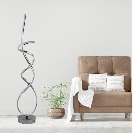 Stojacia lampa s kovovou špirálovitou konštrukciou stojí na tmavej podstavci v miestnosti. Vedľa nej sa nachádza hnedé kreslo s vankúšmi a rastlina v kvetináči.