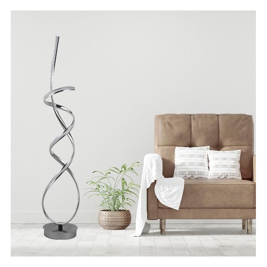Stojacia lampa s kovovou špirálovitou konštrukciou stojí na tmavej podstavci v miestnosti. Vedľa nej sa nachádza hnedé kreslo s vankúšmi a rastlina v kvetináči.