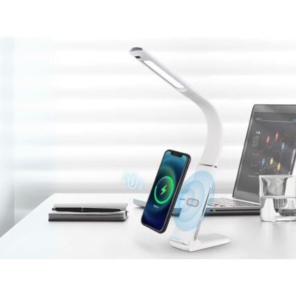 Biela stolová lampa s integrovanou bezdrôtovou nabíjačkou nabíja telefón. Lampa je na stole s notebookom a pohárom vody. Na stole je aj zápisník a pero.