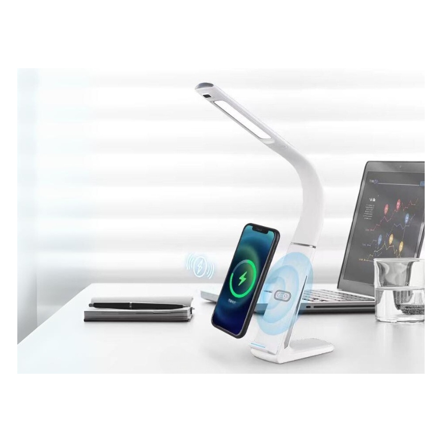 Biela stolová lampa s integrovanou bezdrôtovou nabíjačkou nabíja telefón. Lampa je na stole s notebookom a pohárom vody. Na stole je aj zápisník a pero.