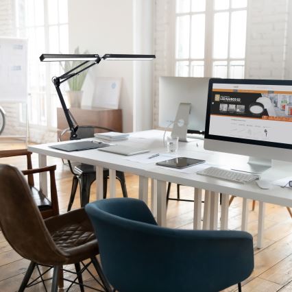 Dvojramenná stolová lampa svieti nad pracovným stolom. Na stole sú počítač, tablet, klávesnica a dokumenty. Prostredie je moderná kancelária. Na monitore počítača je webová stránka s textom „LED NAIROS G2“.