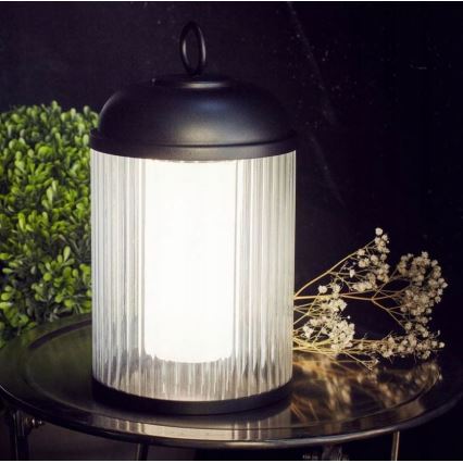 Stolová lampa svieti. Je cylindrická, s čiernym kovový rámom a priehľadným skleneným telom s vertikálnymi ryhami. Stojí na tmavom kovovom podnose, vedľa zelenej rastliny a suchých kvetov.