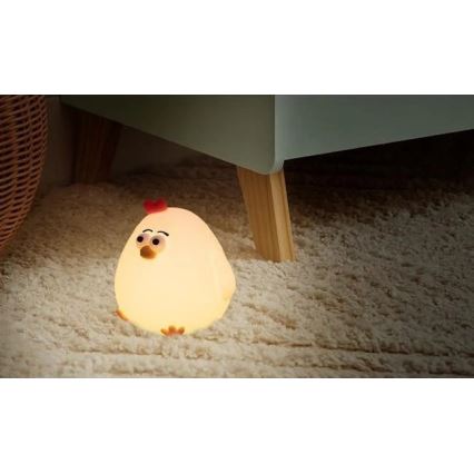 Svietiaca lampička v tvare kuriatka svieti na svetlohnedom koberci pod nábytkom. Kuriatko je svetložlté s červeným hrebienkom. Nábytok má svetlomodrú farbu a drevené nohy.