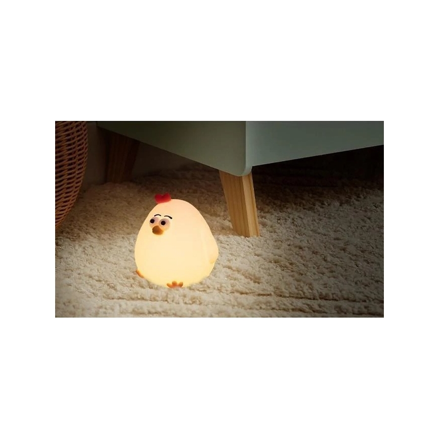 Svietiaca lampička v tvare kuriatka svieti na svetlohnedom koberci pod nábytkom. Kuriatko je svetložlté s červeným hrebienkom. Nábytok má svetlomodrú farbu a drevené nohy.