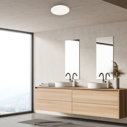 Svietidlo svieti v modernej kúpeľni. Dvojité umývadlo s drevenou skrinkou je umiestnené pod dvoma zrkadlami. Veľké okno poskytuje výhľad do krajiny.