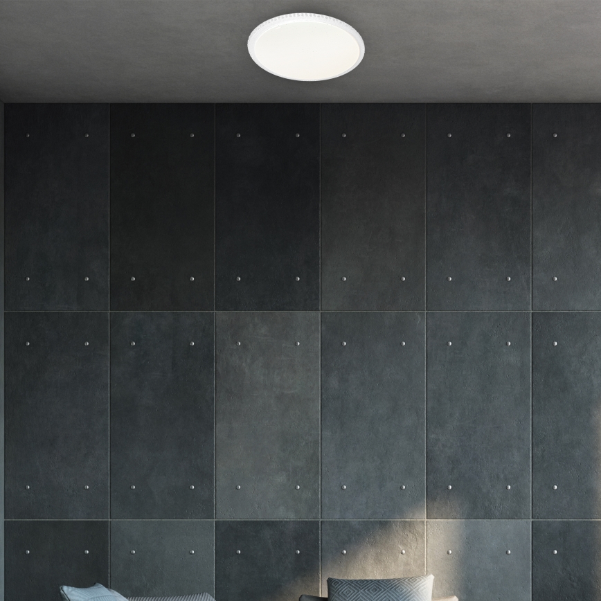 Okrúhle stropné svietidlo svieti v miestnosti s tmavosivou stenou z panelov. Panely sú pripevnené kovovými nýtmi. V popredí sú naznačené vankúše na sedačke.