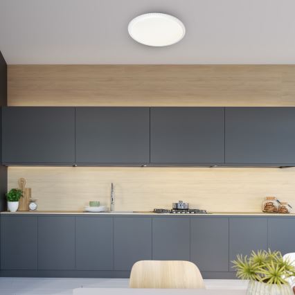 Svetlo svieti v modernej kuchyni. Okrúhle stropné svietidlo je umiestnené nad tmavosivou kuchynskou linkou s drevenou stenou. V kuchyni je viditeľná varná doska a drez.