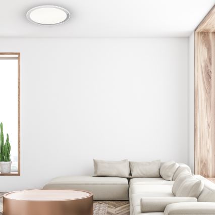 Svetlo béžová rohová sedačka stojí v miestnosti s bielymi stenami a drevenými prvkami. Pred sedacou súpravou sa nachádza nízky okrúhly stolík s medeným povrchom. V pozadí je vidieť okno a kaktus v kvetináči.