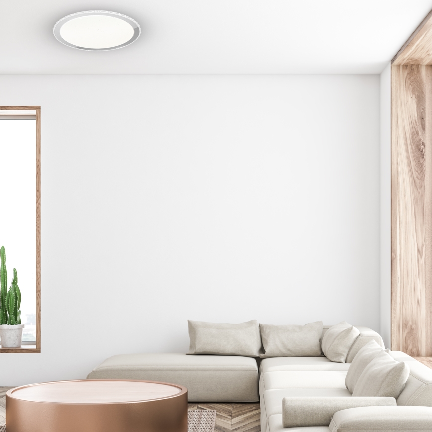 Svetlo béžová rohová sedačka stojí v miestnosti s bielymi stenami a drevenými prvkami. Pred sedacou súpravou sa nachádza nízky okrúhly stolík s medeným povrchom. V pozadí je vidieť okno a kaktus v kvetináči.